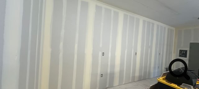 A room with unfinished drywall on the walls, a concrete floor, and some construction materials and tools in the foreground.