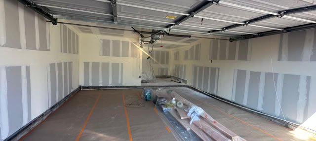 Interior of an unfinished garage with drywall on the walls, a concrete floor covered with protective plastic, and construction materials in the center.