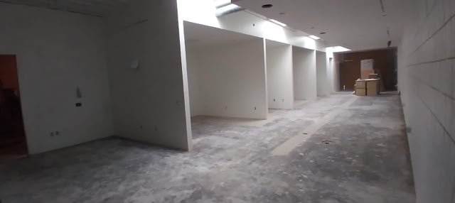 A long, empty hallway with white walls and a concrete floor, featuring several open doorways on the left and stacked boxes at the far end.