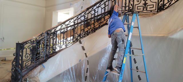 A person on a ladder working on a staircase with ornate metal railings, surrounded by protective plastic sheeting.