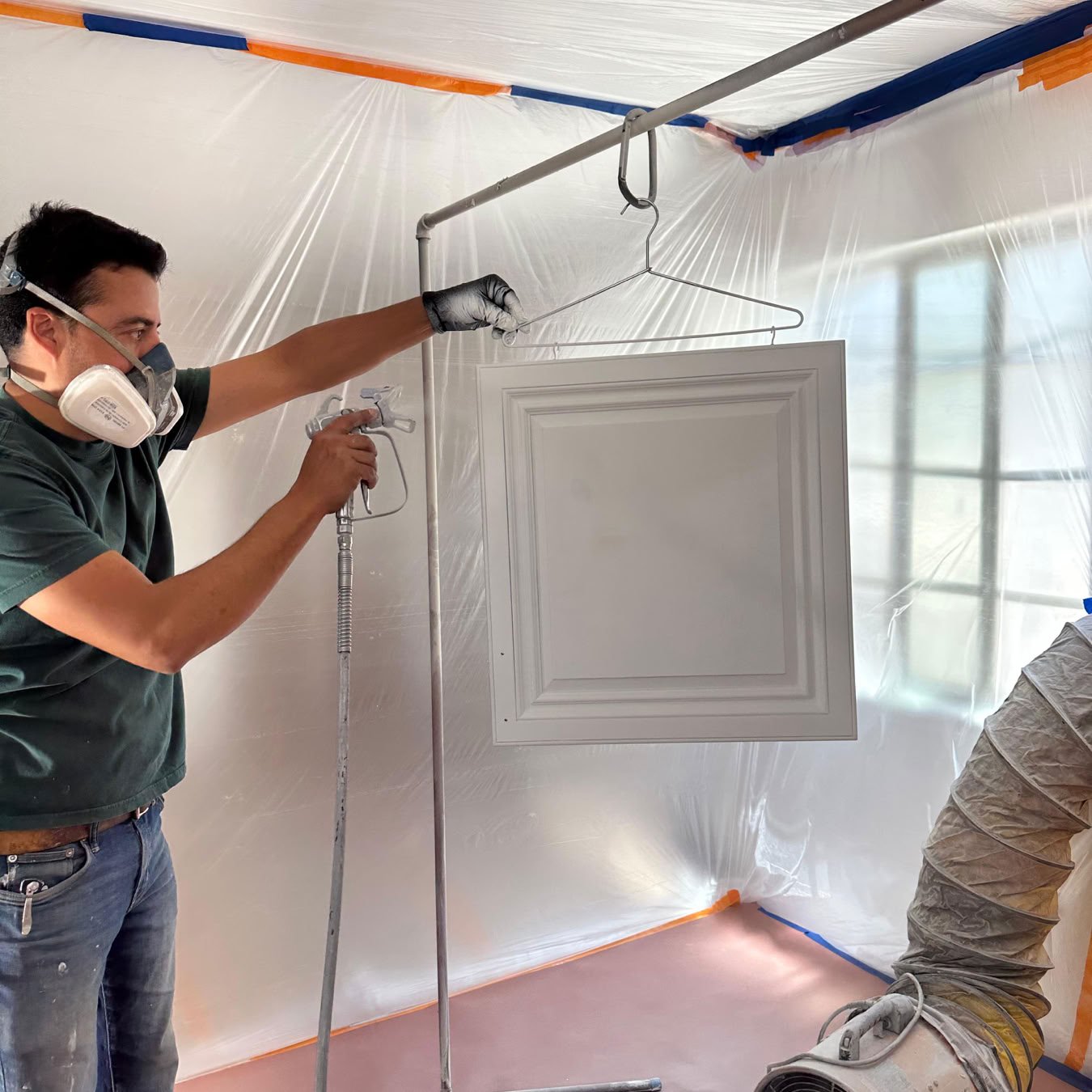 Back view of a painter in overalls and hard hat painting an interior wall with a roller.